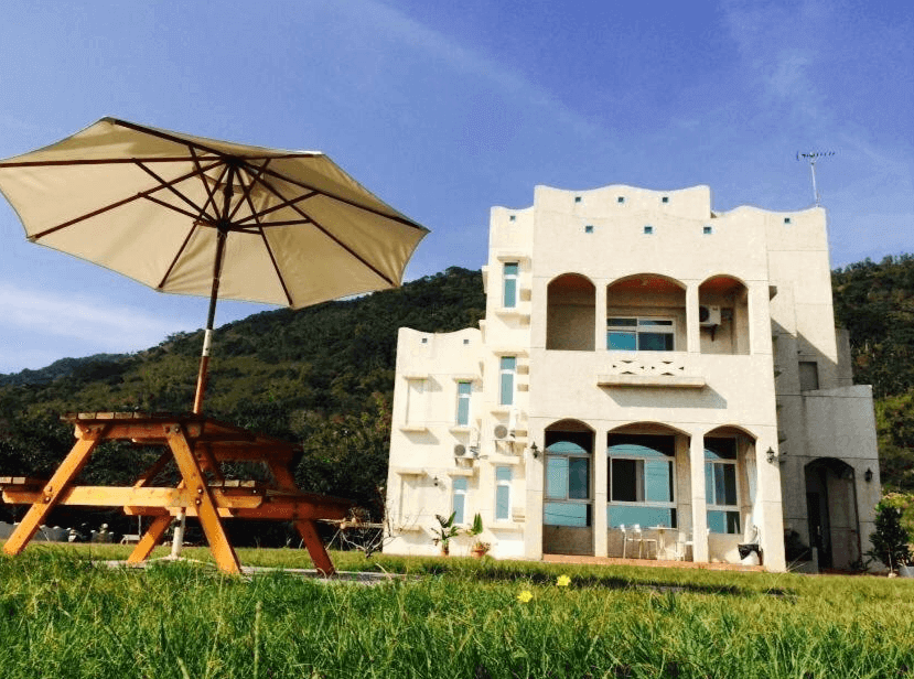 【豐濱住宿】海公主民宿外觀為白色地中海風格建築，前方草地上設有大型陽傘與木製野餐桌椅，坐擁山林與藍天，環境清幽自然，是親近大自然的度假選擇。