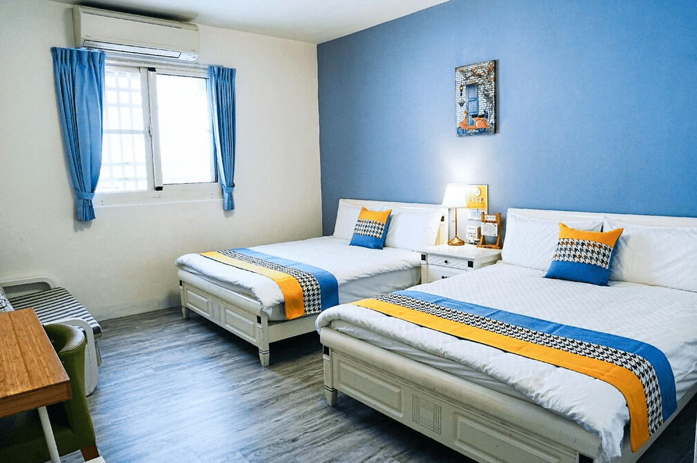 【布袋住宿】海風長堤民宿海洋主題四人房，設有兩張雙人床，牆面採用藍白配色，搭配橘色與藍色跳色床巾與抱枕，整體風格清新明亮，窗邊設有書桌與椅子。
