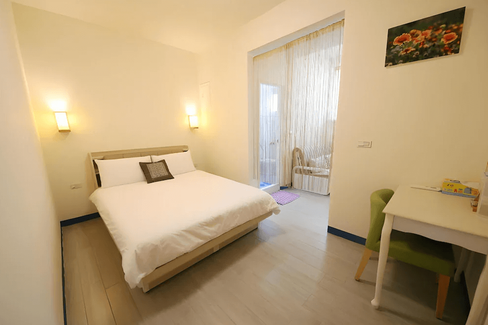 【布袋住宿】海風長堤民宿溫馨雙人房，空間簡潔，設有一張雙人床與白色書桌，牆上裝飾花卉畫作，延伸區域有透明簾幕與藤編搖椅，營造寧靜舒適氛圍。