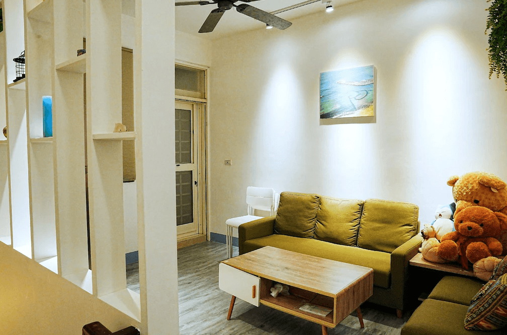 【布袋住宿】海風長堤民宿交誼客廳空間，擺放綠色沙發與木質茶几，角落有可愛玩偶與抱枕，牆上掛有海洋畫作，空間溫馨適合親子共聚或好友聊天。