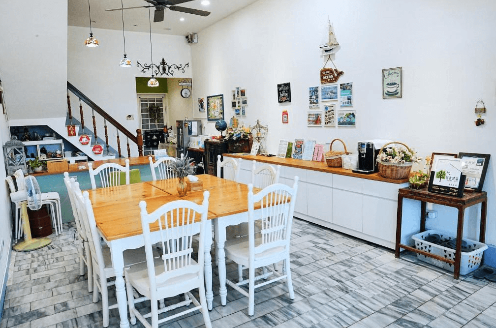 【布袋住宿】海風長堤民宿用餐及公共空間，設有長方形木質餐桌與白色餐椅，牆面裝飾多幅海洋風明信片與小擺飾，旁邊備有咖啡機、旅遊資訊與自助吧台。