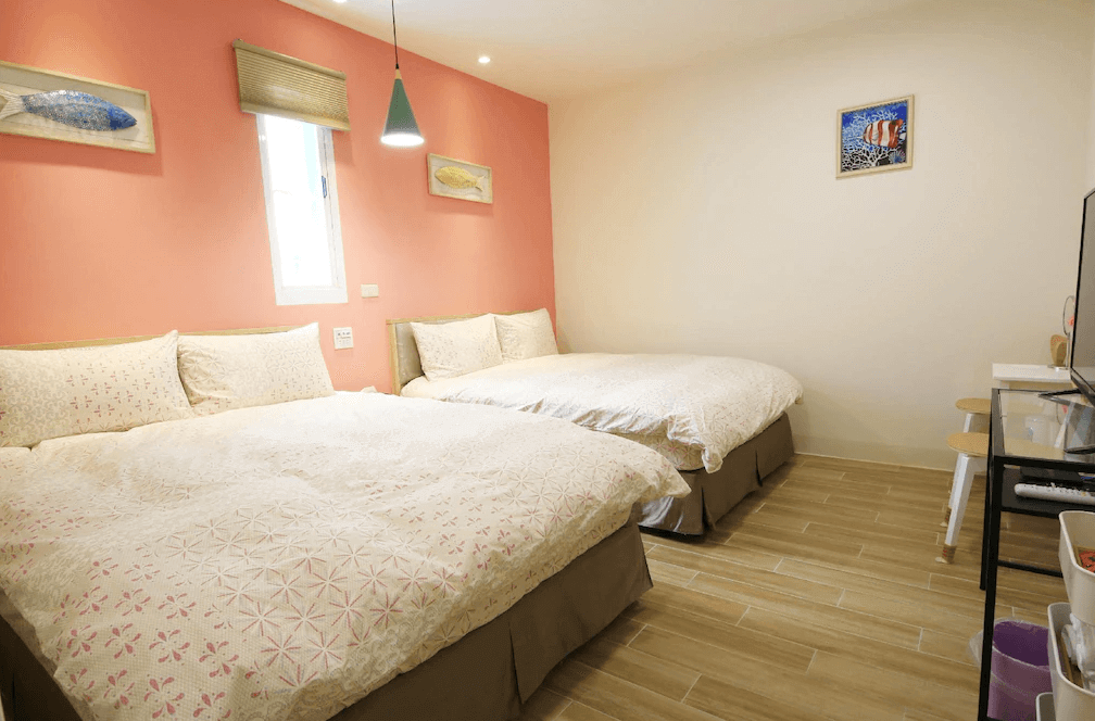 【布袋住宿】海風長堤民宿二館溫馨四人房，房內配置兩張雙人床，牆面為柔和粉紅色，搭配可愛魚類掛畫與木地板，空間舒適，適合親子與好友同行。