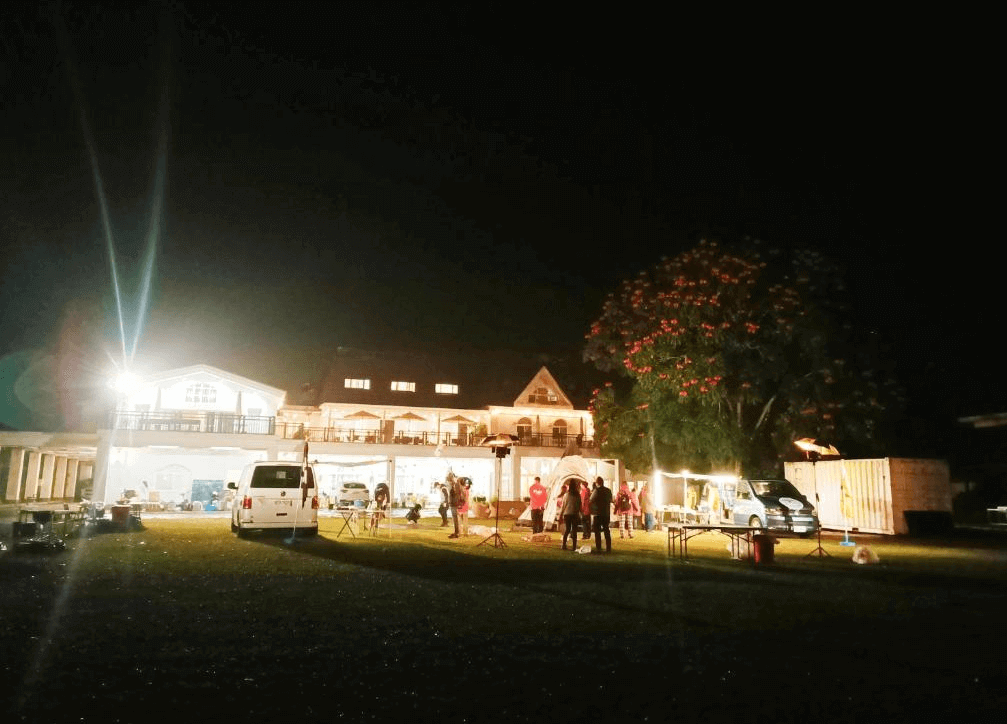 【玉井住宿】瓦樣山巷夜晚拍攝的戶外活動場景，前方草地上搭建帳篷並擺設燈光與設備，人們聚集參加活動，背景是一棟白色雙層洋房建築，屋後有一棵盛開紅花的大樹，營造出溫馨歡樂的露營氣氛。