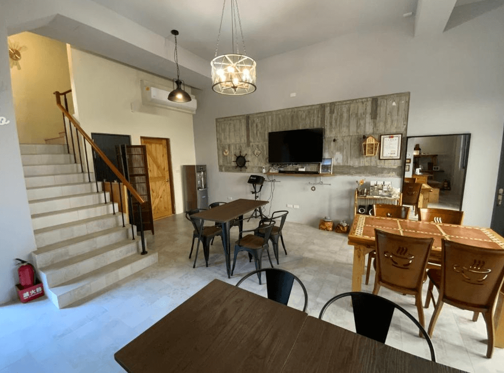 【長濱住宿】米粒教育家一樓公共交誼空間，備有多組木桌椅、電視與展示牆，並設有樓梯通往客房樓層，整體裝潢溫暖簡潔。