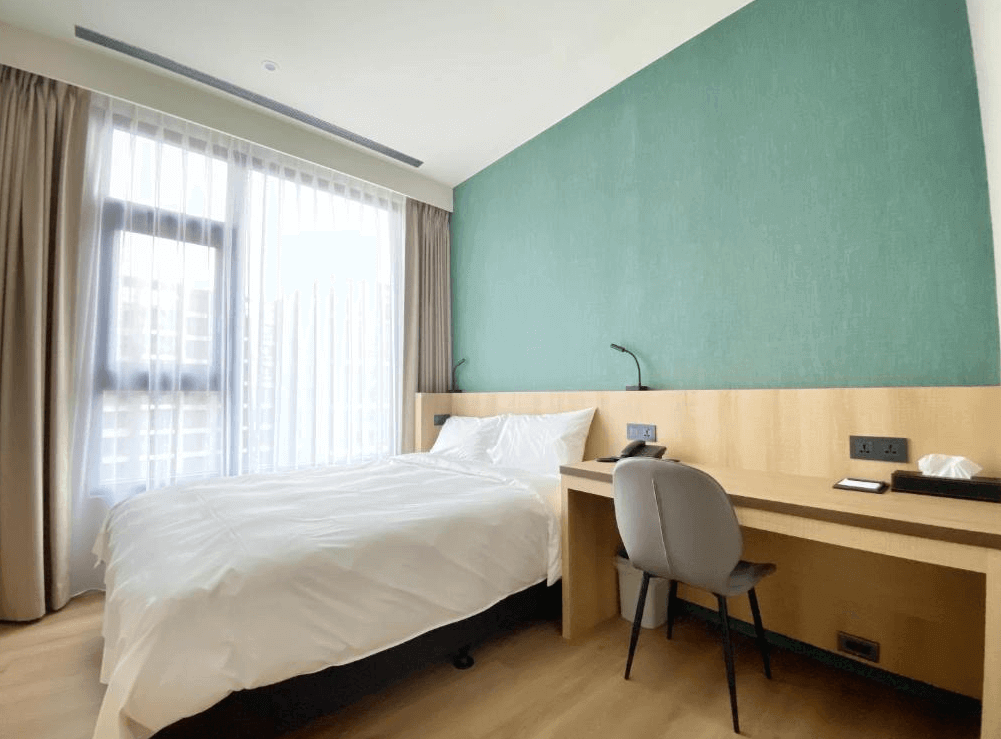 【桃園機場住宿】艾爾芙公園行旅的簡約風格客房，配有書桌、雙人床與柔和綠色牆面，窗簾採用透光設計，空間清新寧靜。