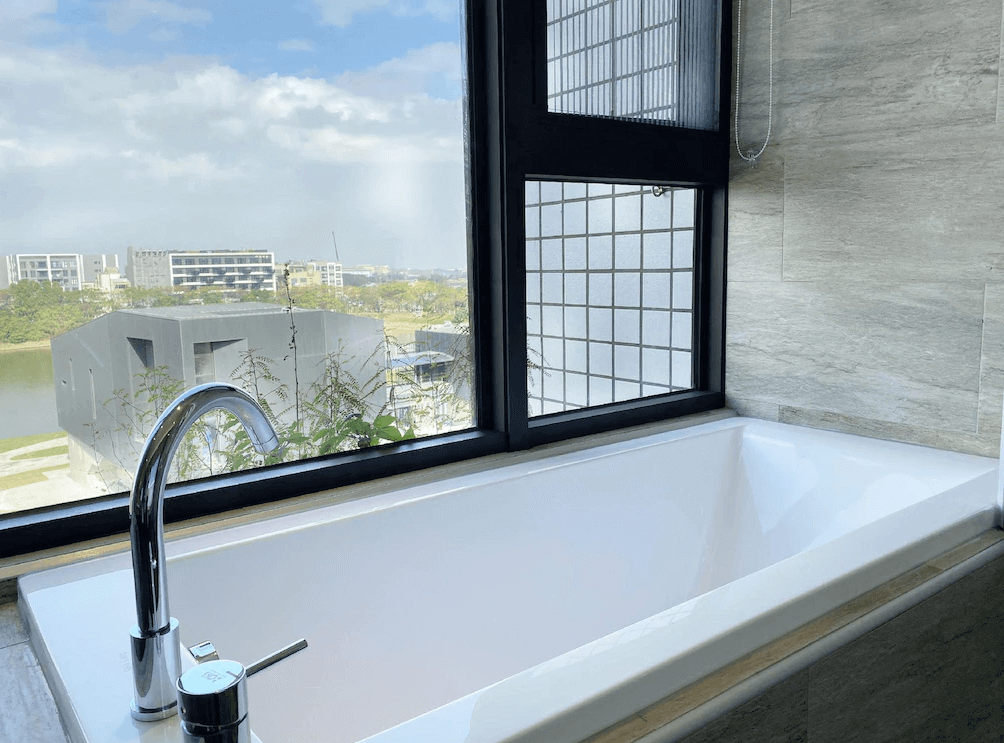 【桃園機場住宿】艾爾芙公園行旅的浴缸景觀窗設計，泡澡同時可眺望戶外綠意與建築群，浴室牆面以淺灰石紋磁磚打造，質感現代。