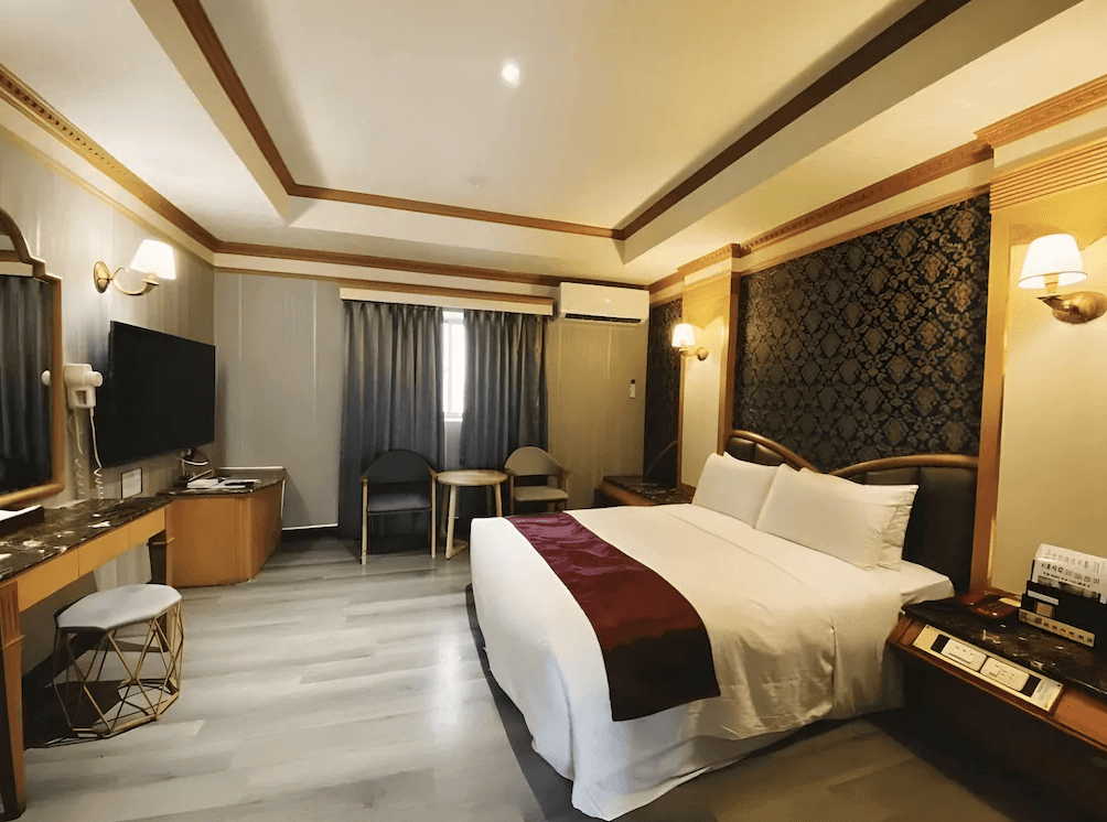 【高雄寵物友善住宿】艾萊商務汽車旅館的雙人房，配有大床、皮革床頭與深色雕花牆面，房內設有梳妝台、電視與小沙發組，整體風格沉穩高雅，空間寬敞舒適。