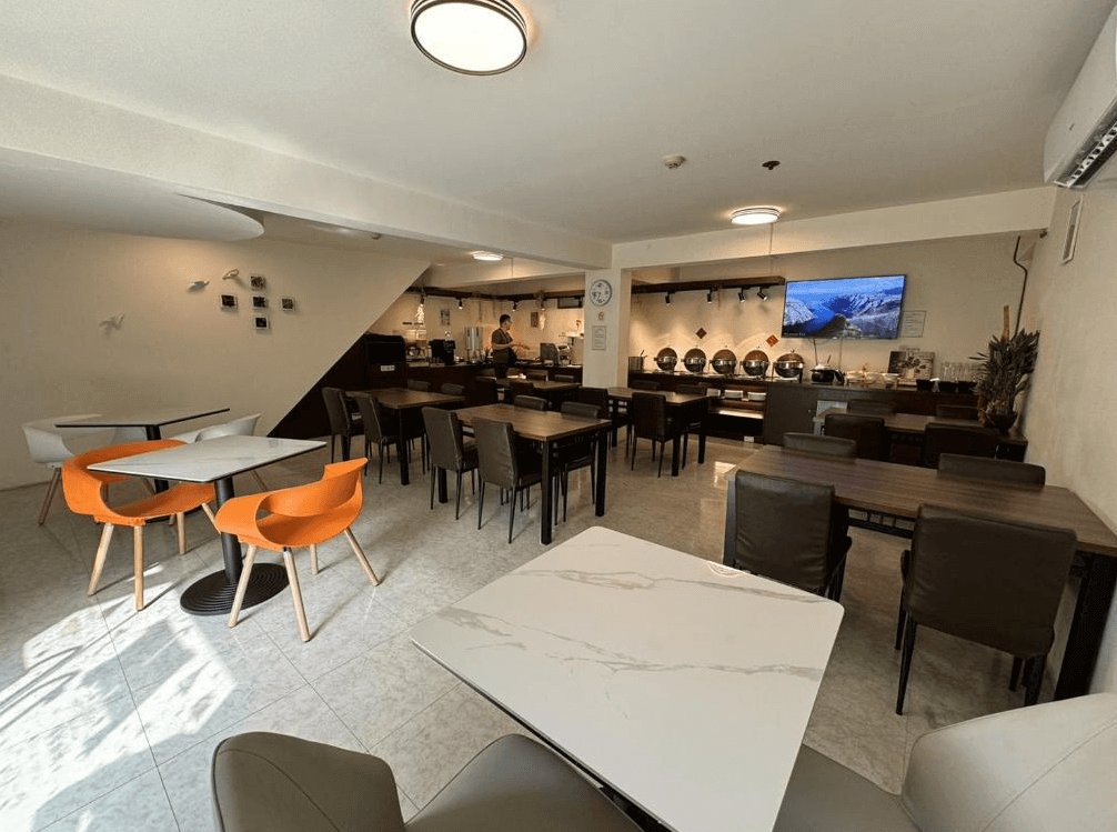 【高雄寵物友善住宿】艾萊商務汽車旅館的用餐區域，設有多組方形餐桌椅，後方為自助式早餐吧檯，並配有電視牆與簡約裝飾，適合旅客早晨用餐與放鬆。