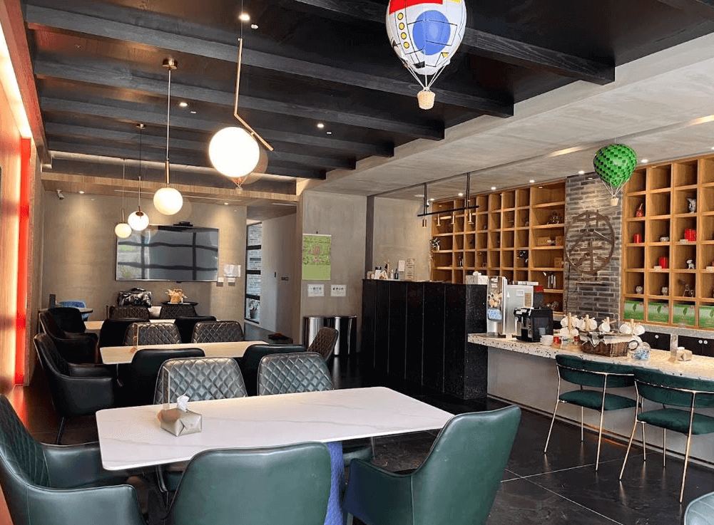 【關山住宿】茶田關舍二館民宿的用餐與接待空間，設有多組皮革座椅與簡約風吊燈，牆面為溫潤水泥質感，後方展示架上擺滿茶具，整體設計雅致又有特色。