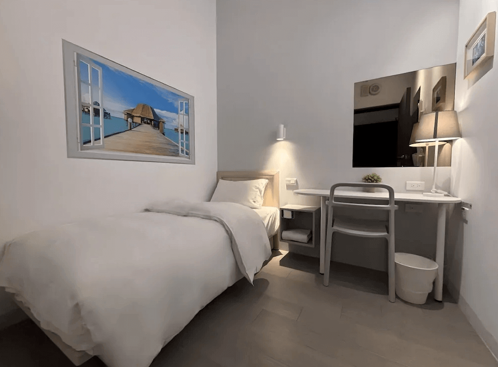 【Xpark住宿】藍水輕旅單人房，房內設有一張單人床、書桌與大面鏡子，牆上掛有度假風格的窗景畫作，整體風格簡約明亮。