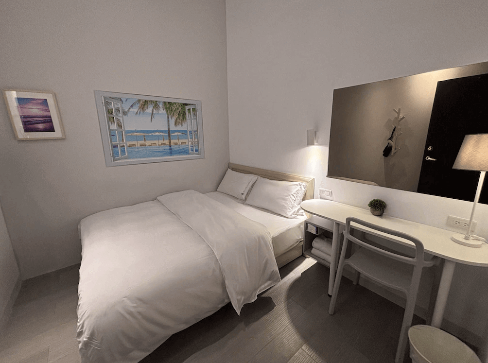 【Xpark住宿】藍水輕旅雙人房，配有舒適雙人床、簡約白色書桌與小盆栽裝飾，牆上掛有熱帶海灘風景畫，營造出輕鬆氛圍。