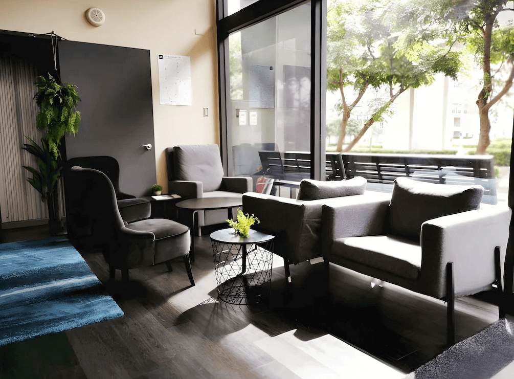 【Xpark住宿】藍水輕旅大廳休息區，設有多組灰色沙發與茶几，大面玻璃窗引入自然光，窗外可見綠意盎然的景致。