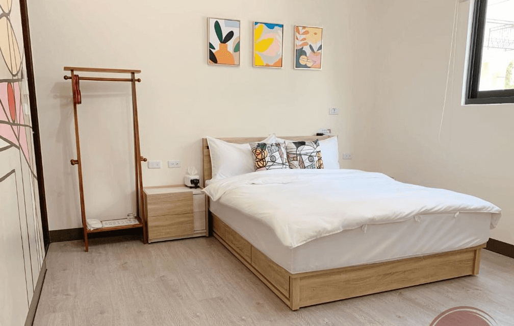 【玉井住宿】藝海民宿的簡約雙人房型，使用自然木質色調搭配白色牆面，床鋪舒適整潔，牆上以三幅色彩鮮明的幾何風格掛畫點綴，設有簡單木質衣架與床頭櫃，整體氛圍明亮溫馨。