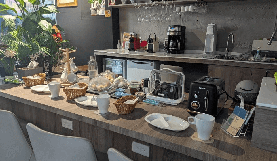 【高雄寵物友善住宿】蘆思特六合民宿的吧台上擺放多樣早餐備品，包括烤麵包機、咖啡機、茶杯、吐司與果醬等，周圍佈置植物與杯架，營造溫馨又便利的用餐空間。