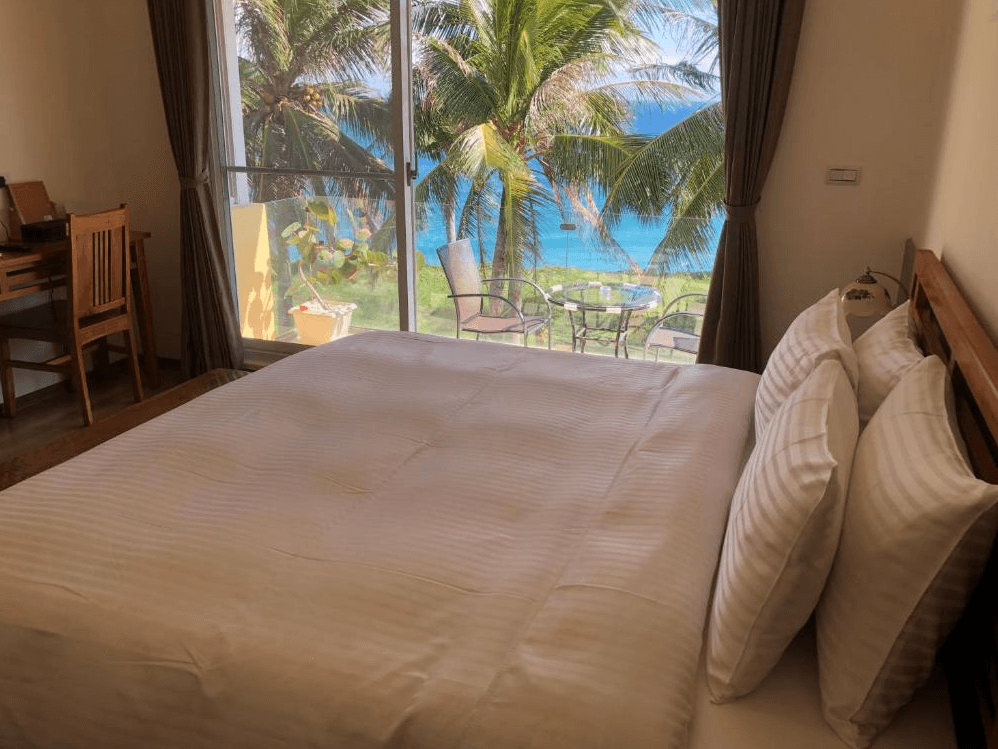 【東河住宿】蘭花楹海景民宿雙人房，房間內鋪設白色床單，落地窗外即是海景陽台，椰子樹與蔚藍海洋一覽無遺，配有木製書桌與座椅，自然採光充足，充滿度假氛圍。