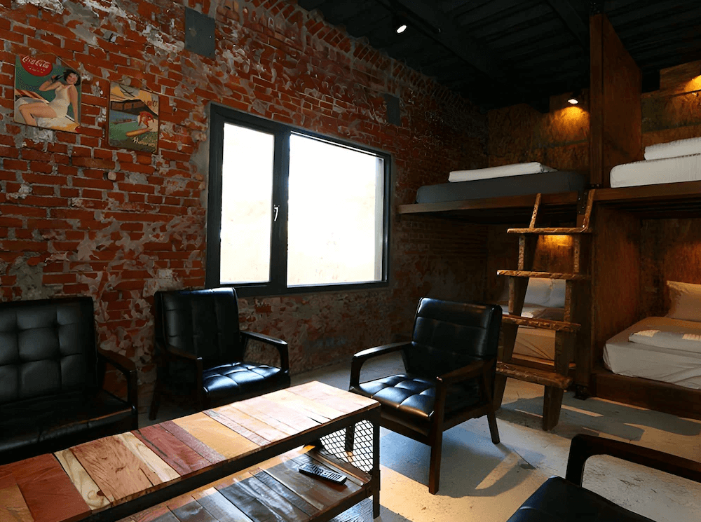 【東河住宿】螺絲釘民宿工業風多人房，牆面為裸露紅磚搭配復古海報，房內設有上下舖木床與皮革沙發座椅，昏黃燈光營造復古與粗獷兼具的風格。