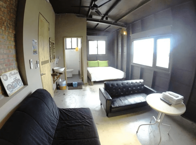 【東河住宿】螺絲釘民宿工業風雙人套房，配置雙人床、沙發、圓桌與開放式衛浴空間，牆面為金屬灰與木質混搭，地面為水泥質感，展現自由不羈的設計風格。