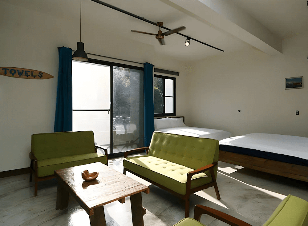 【東河住宿】螺絲釘民宿四人房，設有兩張雙人床與淺綠色復古沙發組，大片落地窗引入陽光，室內採用木質家具與吊扇，空間溫馨寬敞，適合家庭與朋友共住。