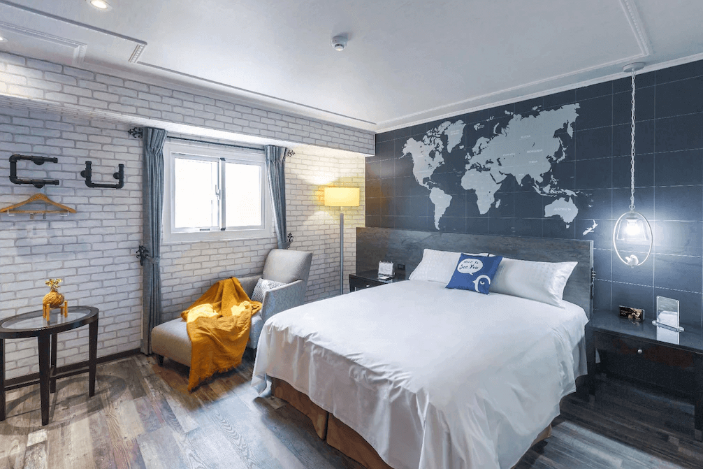 【高雄寵物友善住宿】西悠巢旅-高雄車站館的房內以灰藍色世界地圖為主題牆，搭配白色磚牆與木質地板，呈現出輕工業與旅人風格，配有雙人床、舒適單椅與暖色調毛毯，整體氛圍溫馨且具設計感。