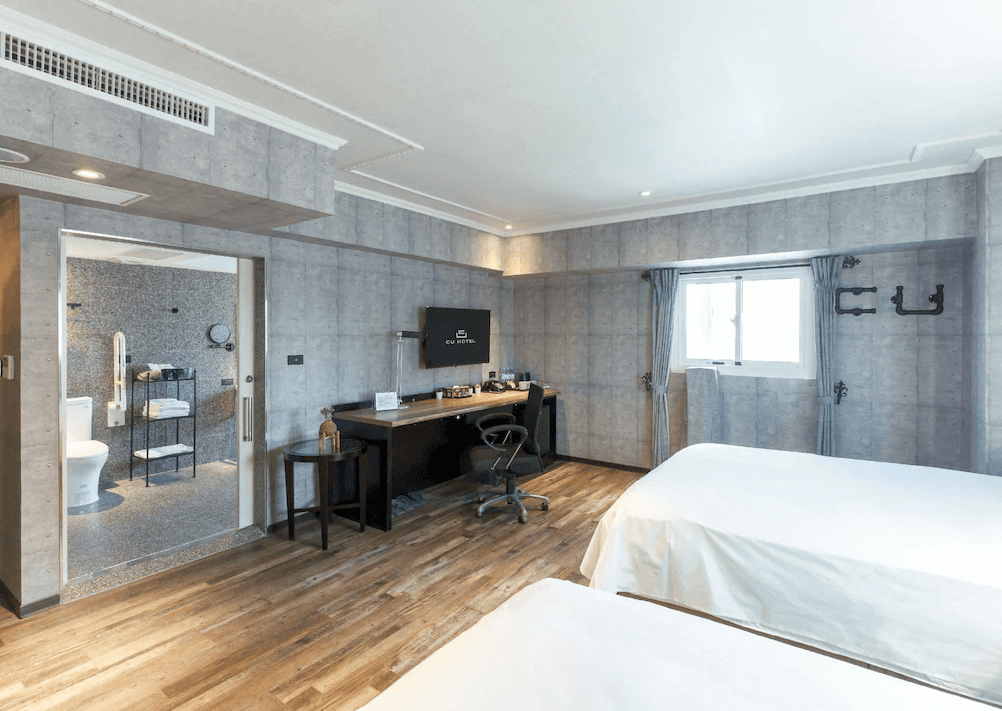 【高雄寵物友善住宿】西悠巢旅-高雄車站館的房內為雙床設計，牆面以灰色仿混凝土風格呈現，搭配木地板與簡約窗簾，空間明亮乾淨，並設有辦公桌與平面電視，開放式衛浴設計帶來便利與視覺延伸感。