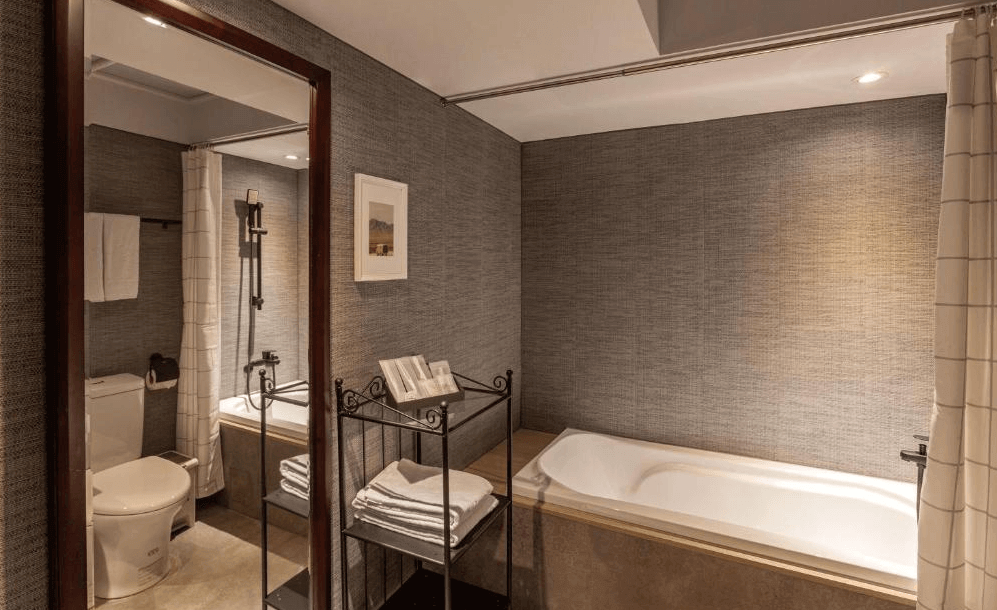【高雄寵物友善住宿】西悠巢旅-高雄車站館，採用深灰紋理牆面與暖黃燈光，設有浴缸與淋浴空間，配置黑鐵毛巾架與鏡面，整體設計簡潔高雅，適合放鬆泡澡與享受私密時光。