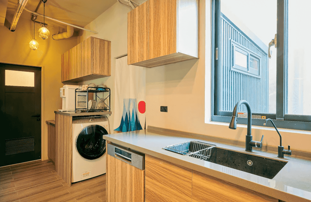 【Xpark住宿】蹤遊三四五共用廚房空間，配有洗衣機、烘碗機、流理臺與烘碗架，並設有窗戶與吊燈，環境整潔溫馨，適合長住旅客使用。