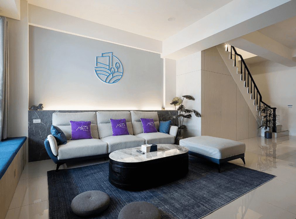【玉井住宿】鈺景 NU TIME 的公共客廳空間，設計感十足，設有大型灰色沙發與圓形大理石茶几，牆面裝飾有住宿 LOGO，搭配深藍色地毯與樓梯旁的植物造景，展現現代簡約與舒適的交融。