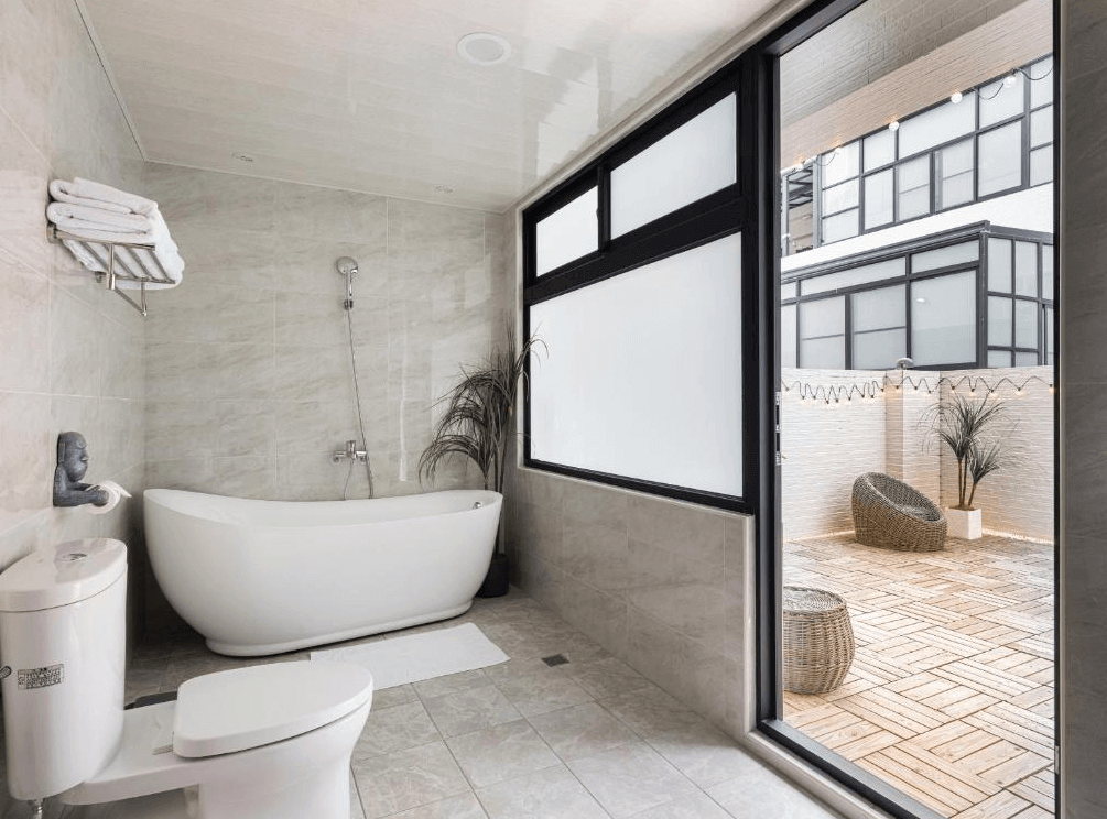 【玉井住宿】鈺景 NU TIME 的浴室空間，設有獨立白色浴缸與馬桶、乾濕分離淋浴設計，大片窗戶與玻璃門通往戶外露台區，露台鋪設木質地板並設有戶外坐墊與綠植，營造度假般的放鬆氛圍。