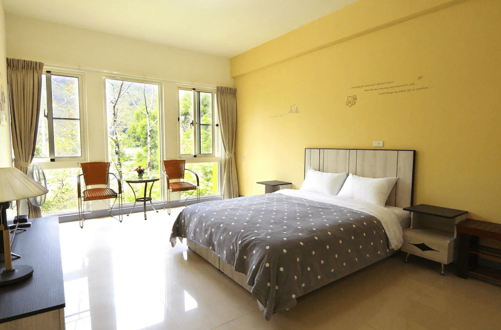 【苗栗包棟民宿】麥克尼景觀莊園的雙人房型，配有大面窗戶可欣賞窗外綠意山景，室內以柔和黃色牆面搭配灰白色系床鋪，營造舒適放鬆的住宿氛圍。