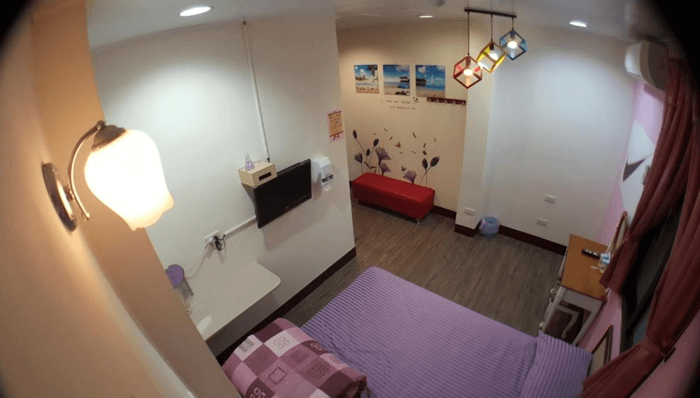 【布袋住宿】龍園旅館雙人房，設有一張雙人床與紫色床單，房內配有壁掛電視、紅色沙發椅、小型書桌與裝飾燈，牆面有海灘風格掛畫與花草牆貼，氣氛溫馨可愛。