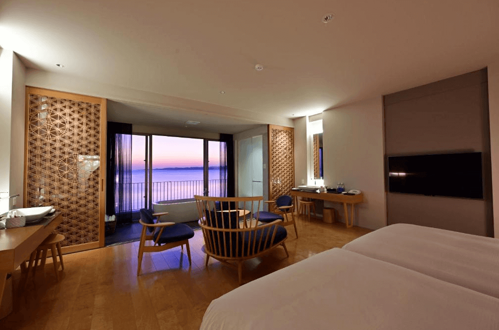【石垣島住宿】Beach Hotel Sunshine 和風海景客房，設有雙人床與日式圓桌座椅，大片落地窗可欣賞夕陽與海景，營造寧靜舒適的住宿氛圍。