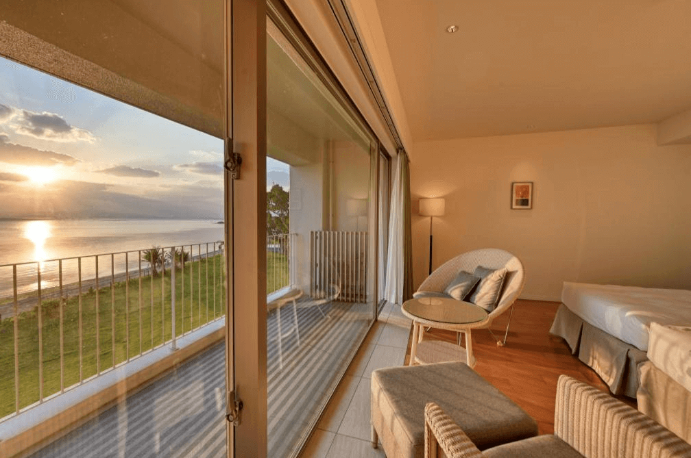 【石垣島住宿】Beach Hotel Sunshine 洋風陽台客房，設有簡約沙發椅與雙人床，透過大片玻璃窗可眺望海岸線與夕陽美景，適合情侶悠閒度假。