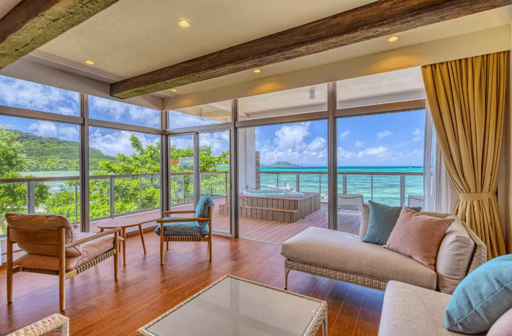 【石垣島住宿】Ishigaki Seaside Hotel 套房客廳空間，設有大片玻璃窗與木質地板，窗外陽台配備私人泡湯池，可眺望蔚藍大海與翠綠山景，充滿渡假感。
