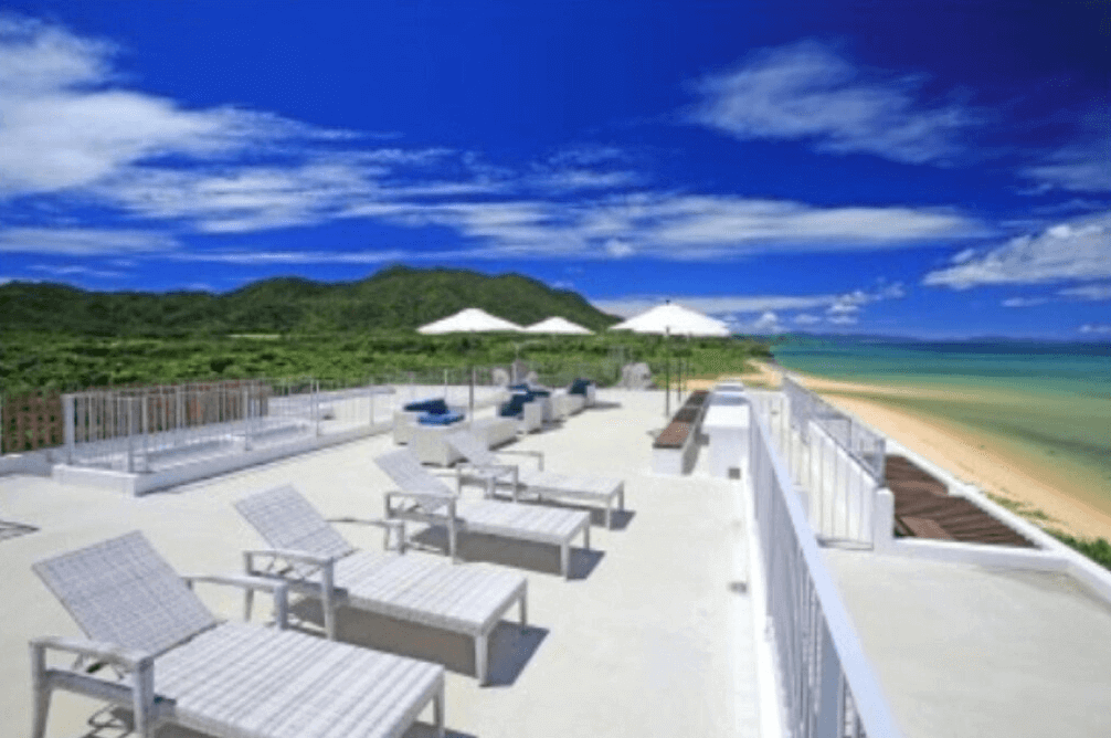 【石垣島住宿】Seven Colors 石垣島海灘景觀平台，設有白色躺椅與遮陽傘，可遠眺翠綠山景與湛藍海洋，為放鬆日光浴的理想空間。