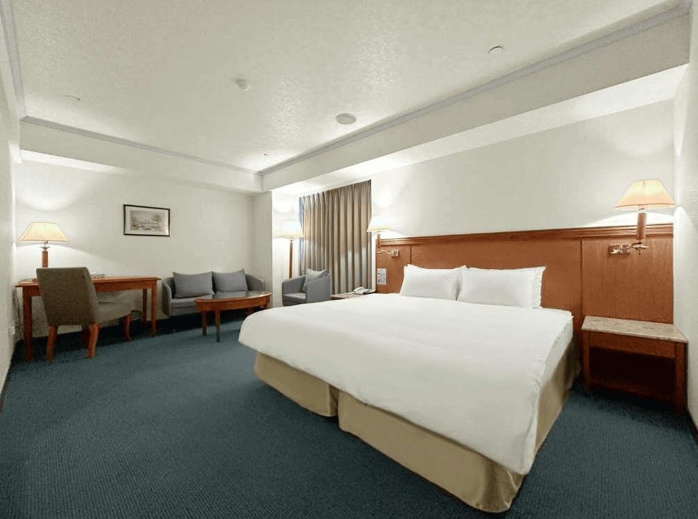 【新竹東區住宿】大道城飯店雙人房空間寬敞，設有一張雙人床、沙發區與書桌，整體風格典雅沉穩，採用柔和燈光營造舒適氛圍。