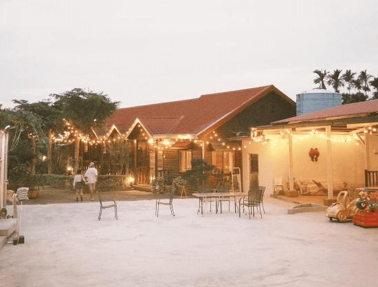 【古坑住宿】天秀山莊餐廳民宿的夜晚戶外空間，主建築屋頂為紅瓦造型，搭配滿布燈串的庭院，擺有休閒桌椅與玩具車，洋溢溫馨與童趣氛圍。