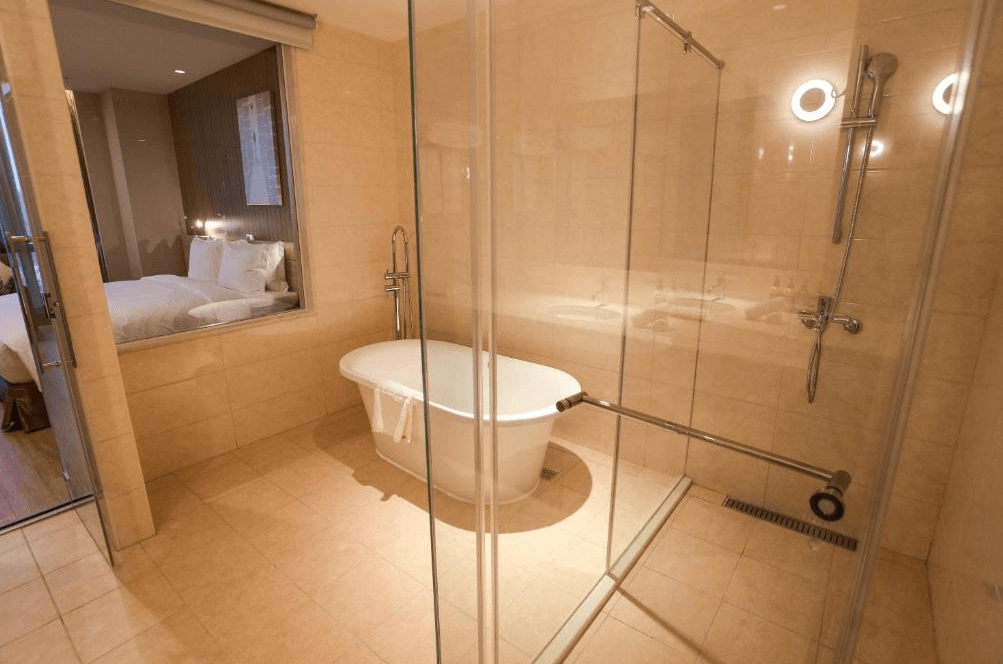 【竹北住宿】新竹安捷國際酒店現代化浴室空間，設有獨立浴缸與玻璃淋浴間，米色磁磚營造溫暖質感，與臥房採透明隔間設計。