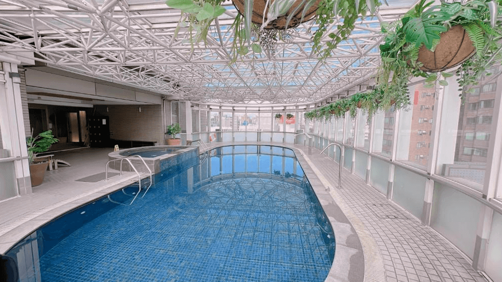 【新竹東區住宿】新竹老爺酒店室內泳池區，設於玻璃帷幕圍繞的溫室空間，天花板採金屬骨架結構並懸掛綠色植栽，環境通透明亮。