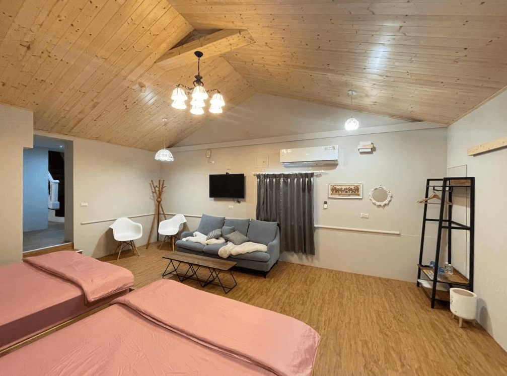 【西螺住宿】旅人路過時親子家庭房全景，兩張雙人床搭配粉色床單與玩偶，整體空間以木質天花板與暖色系地板營造溫暖氛圍，附設沙發與電視區，適合多人入住。