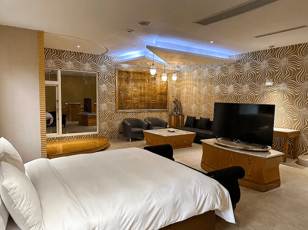 【竹山住宿】歐遊國際連鎖精品旅館竹山館寬敞豪華房型，設有大尺寸雙人床、現代風格沙發組、圓形鏡面泡澡浴池與大螢幕電視，空間裝潢帶有金色幾何牆面與燈飾，展現時尚與舒適並重的氛圍。