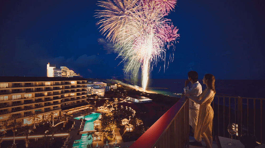 【石垣島住宿】石垣全日空洲際渡假村陽台夜景，一對情侶欣賞著綻放的繽紛煙火，俯瞰整個度假村與泳池區的燈火景致。