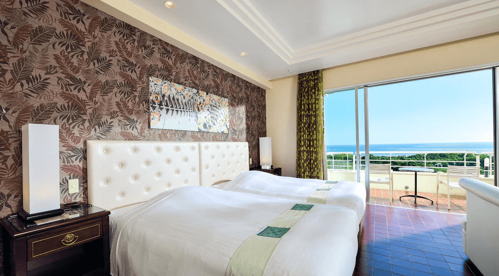 【石垣島住宿】石垣島艾克斯飯店海景雙床房，牆面為熱帶葉紋設計，搭配白色軟包床頭與陽台落地窗，可眺望綠意與遼闊海景，室內風格典雅大方。