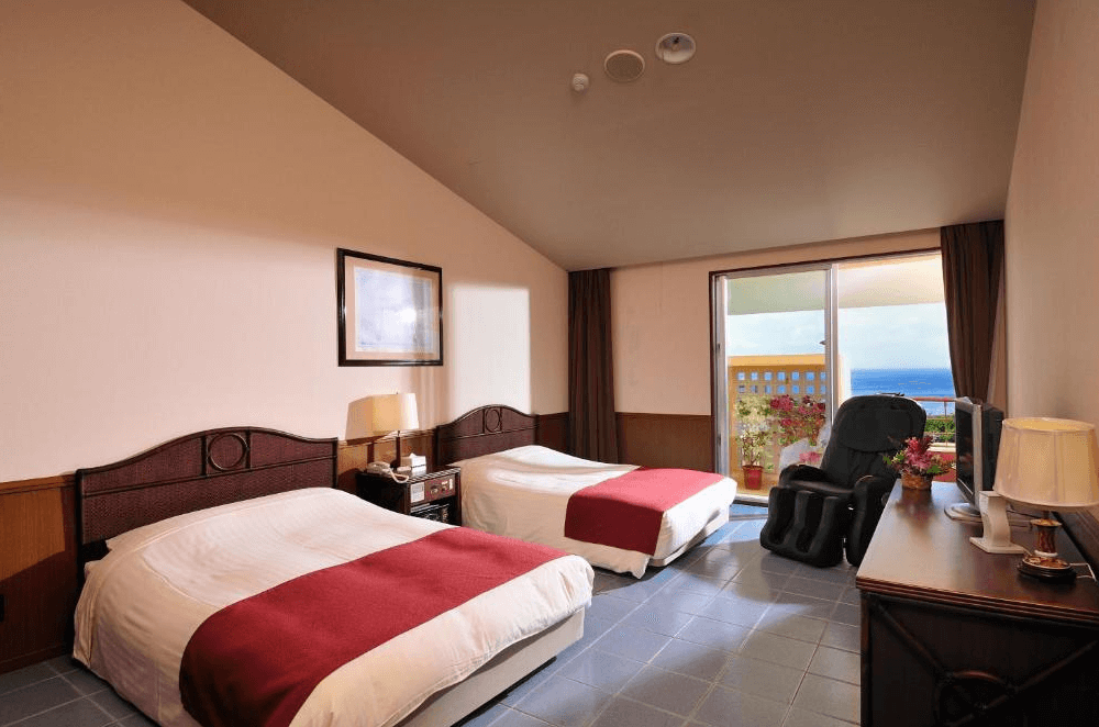 【石垣島住宿】石垣島艾克斯飯店標準雙床房，設有紅色床旗與木質傢俱，房內配有電視與按摩椅，陽台可遠眺海景，氛圍溫馨舒適。