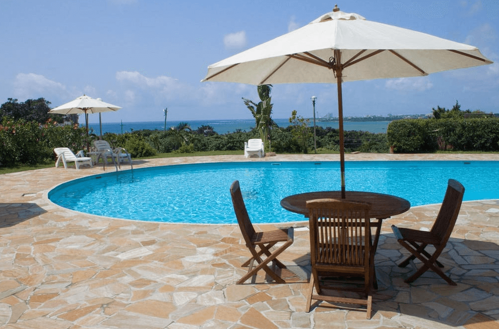 【石垣島住宿】石垣島艾克斯飯店戶外泳池區，設有圓形泳池與木質桌椅、遮陽傘，背景為碧藍海景與綠意植栽，是度假放鬆的理想空間。