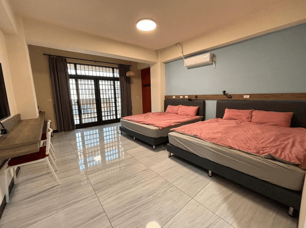 【西螺住宿】粨倉文旅雙人雙床客房，房內擺放兩張大床，搭配粉色寢具，牆面藍色與木質設計，地板為大理石磁磚，設有書桌與椅子，空間寬敞明亮，附有大面落地窗與陽台。