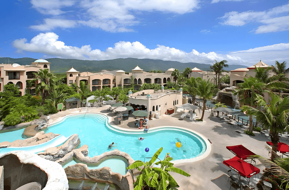 【花蓮寵物友善飯店】俯瞰花蓮理想大地渡假飯店的戶外泳池與園區建築，設施包含滑水道、造型泳池與遮陽休憩區，背景為藍天白雲與中央山脈，展現熱帶渡假村風情。