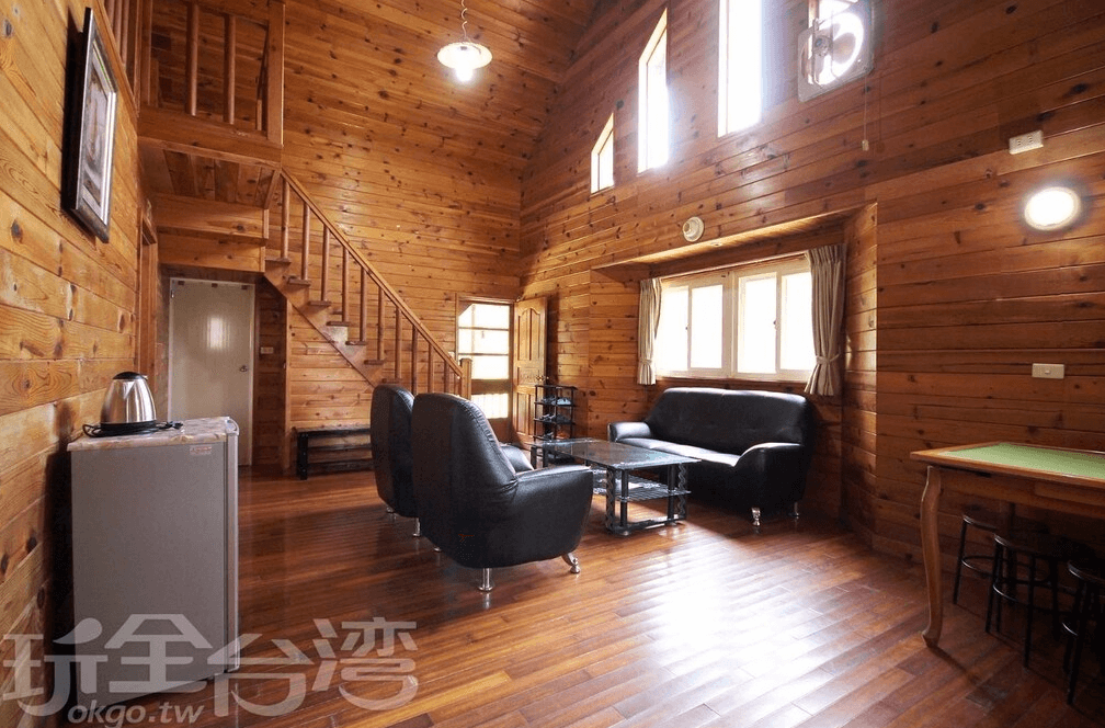 【竹山住宿】雙湖渡假山莊木屋客廳空間，挑高屋頂設計，配有黑色皮沙發與玻璃茶几，木質牆面營造溫馨氛圍，旁邊有樓梯通往二樓。