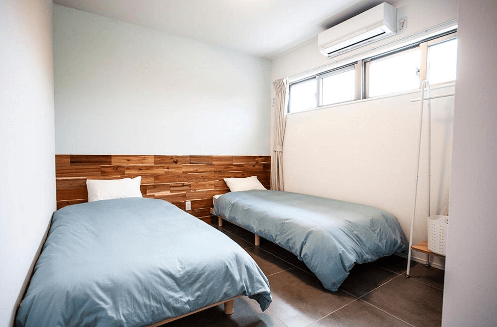 【名護住宿】Hichu House 現代簡約雙床房，木質床頭搭配兩張單人床，淺藍色被套與白色牆面營造清爽氛圍，窗邊設有冷氣與自然採光。