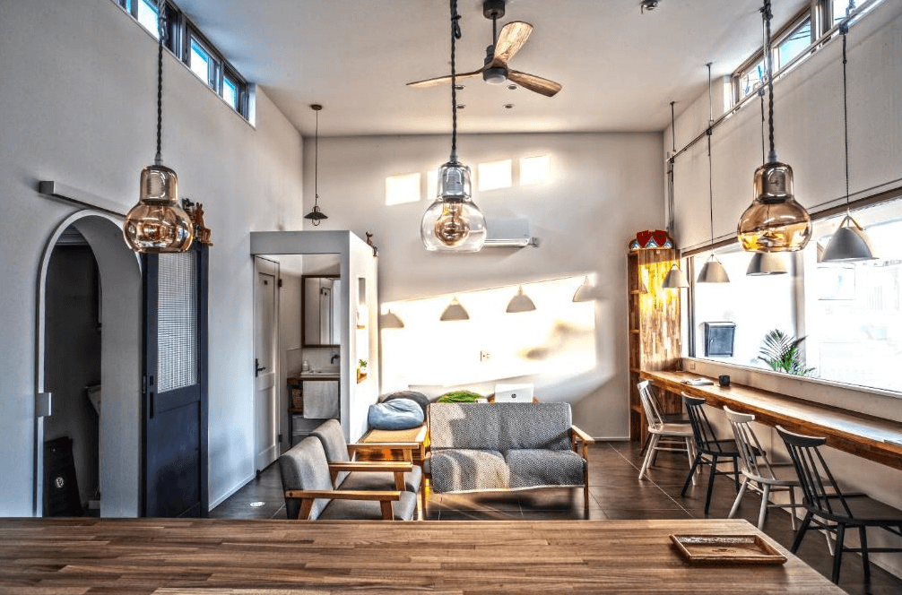 【名護住宿】Hichu House 公共交誼廳，挑高設計搭配吊燈與大片窗戶，自然光灑落，空間內設有沙發、閱讀桌椅與舒適座位區，氛圍溫馨。