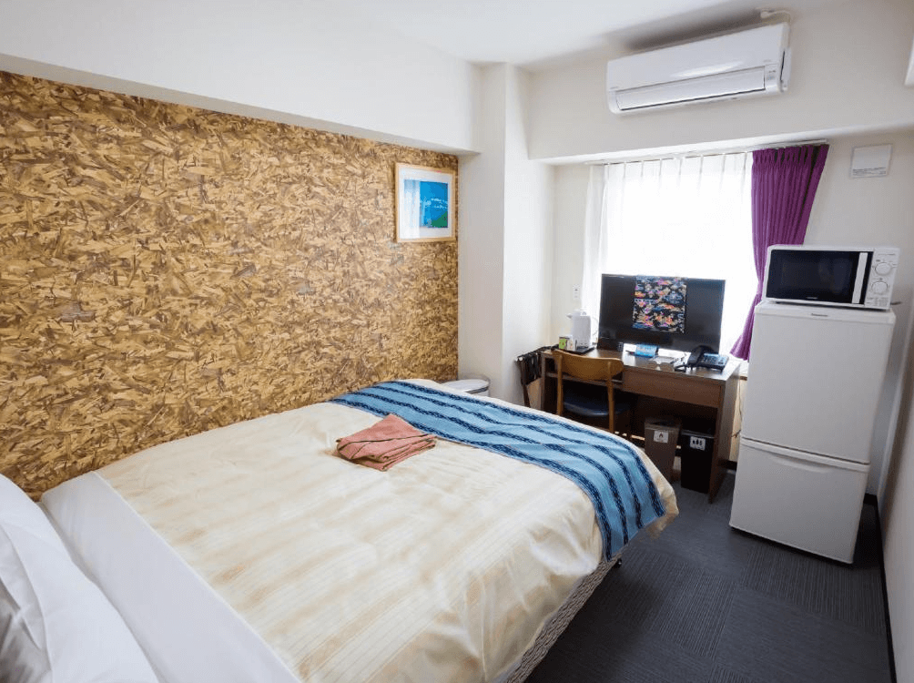 【名護住宿】Hotel Peace Island Nago 雙人房，房內設有大床、書桌、電視與冰箱，牆面採木紋裝飾，空間簡潔實用。